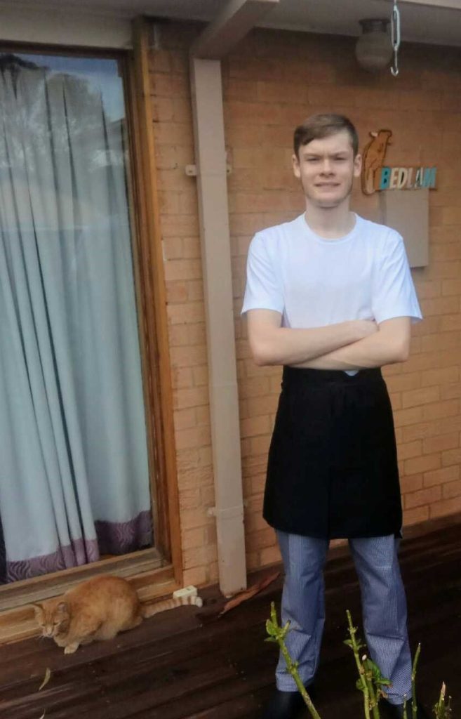 This is a photo of Austin, on his first day of his apprentiship as a chef. He is standing in front of a brick wall, with his arms crossed and smiling. He’s dressed in his chef uniform and has a black apron around his waist. His cat is sitting behind him.