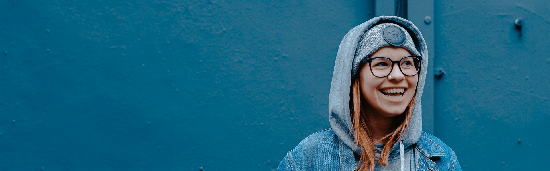 Young woman with a nose ring, wearing a hoodie and glasses in front of an industrial blue wall